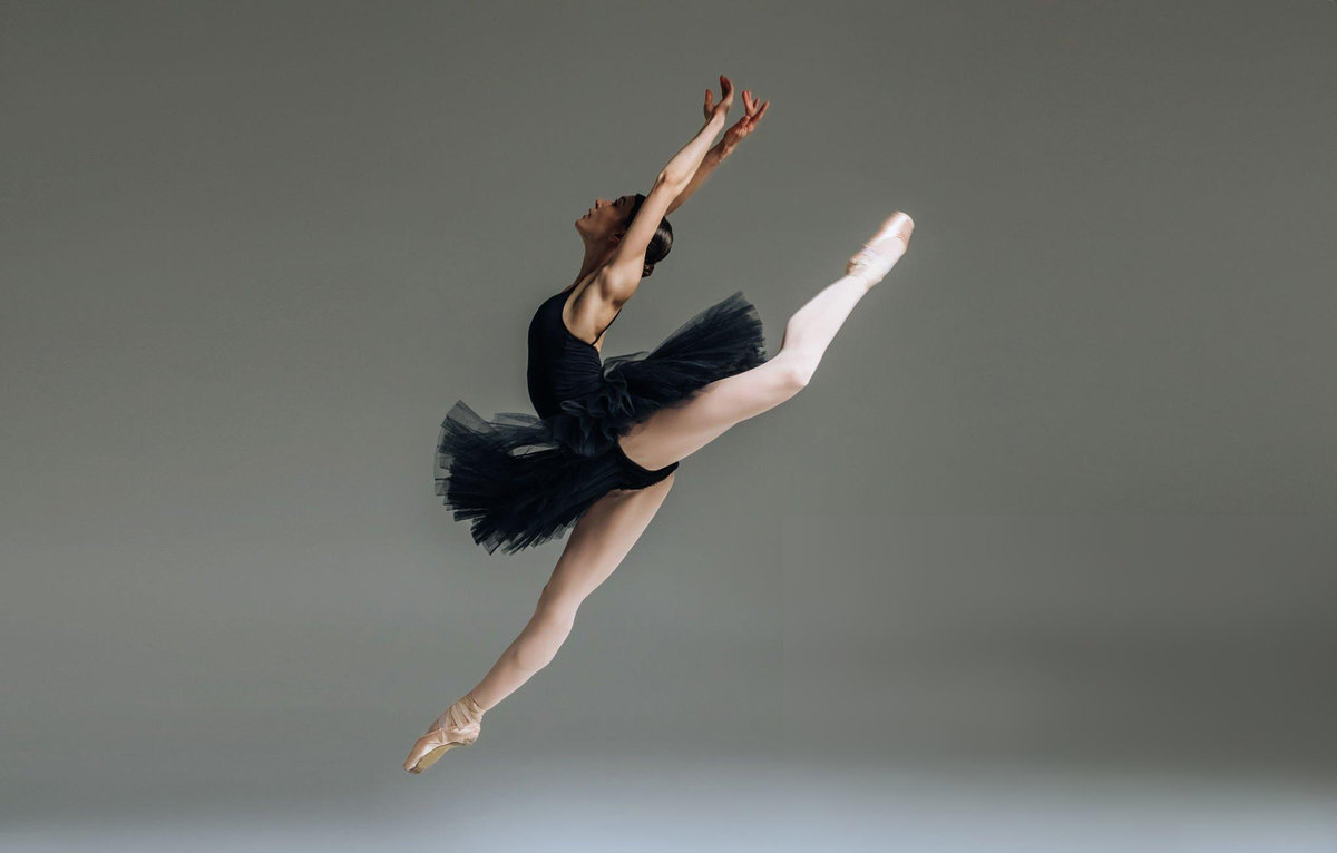 Professional ballerina performing grand jeté leap in black classical tutu and pointe shoes against grey background