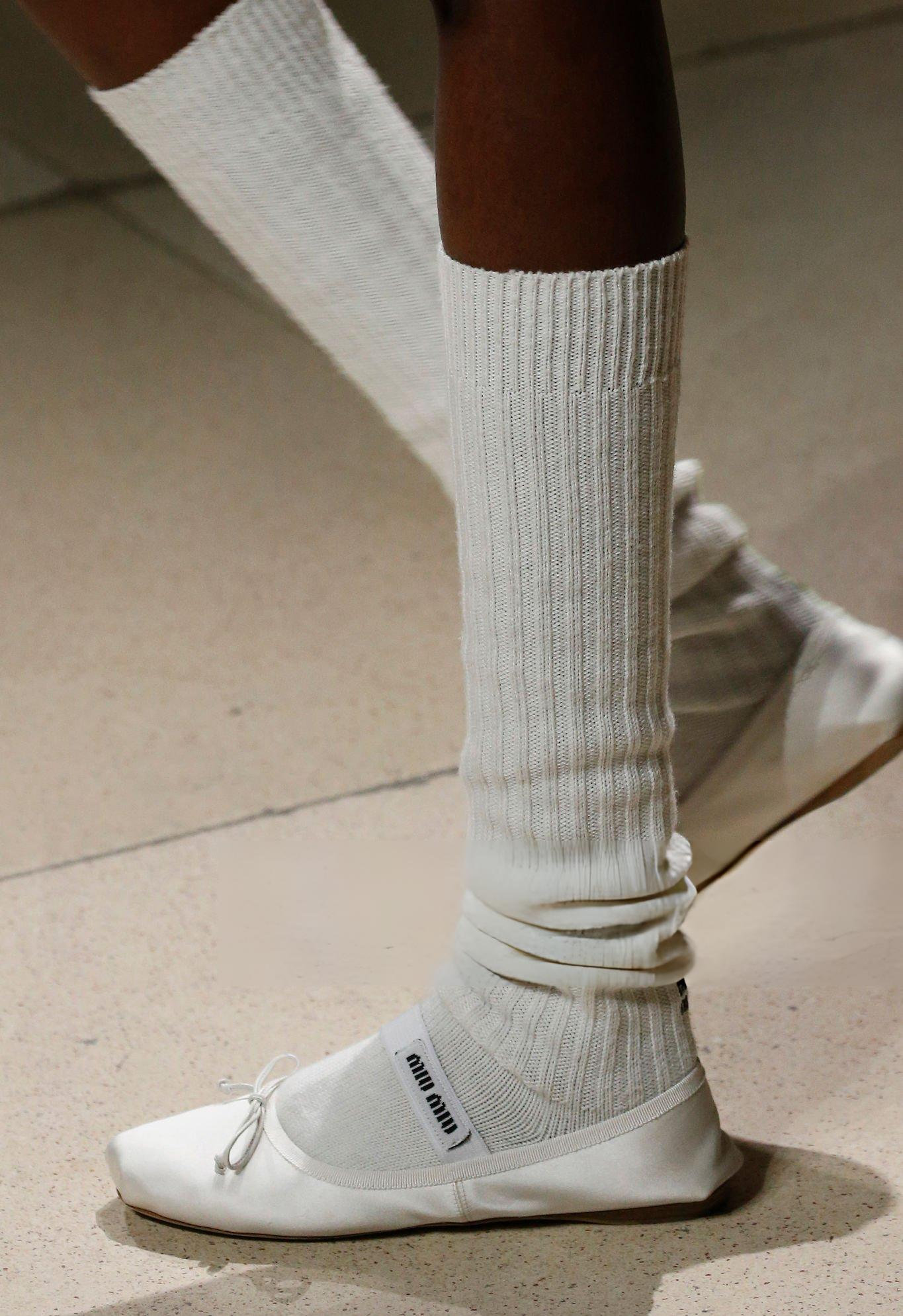 Close-up of Miu Miu Autumn/Winter 2022 white ballet flats with logo elastic strap styled with chunky ribbed white socks on runway