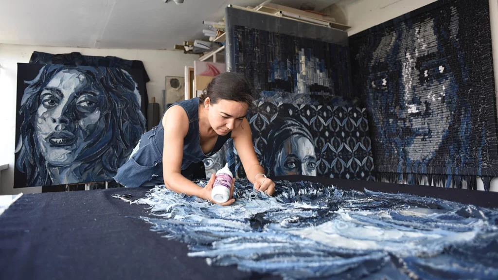 Deniz Sağdıç trabalha meticulosamente em uma peça de arte em jeans em seu estúdio, com retratos completos ao fundo.