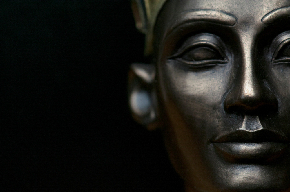 Close de uma escultura que lembra a Rainha Nefertiti do Antigo Egito, com uma profundidade de campo rasa focando seus traços faciais serenos contra um fundo escuro.