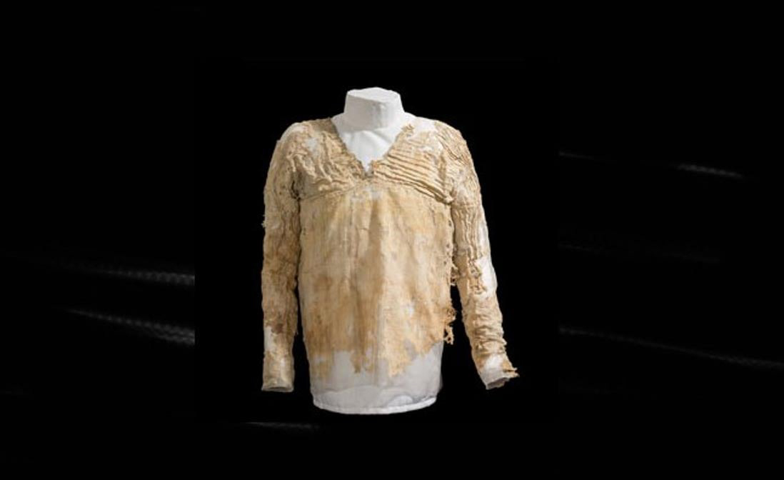 The Tarkhan Dress, an ancient Egyptian garment displayed on a mannequin, featuring a delicate beige lace-trimmed blouse with a V-shaped neckline and long, flowing sleeves against a dark background.