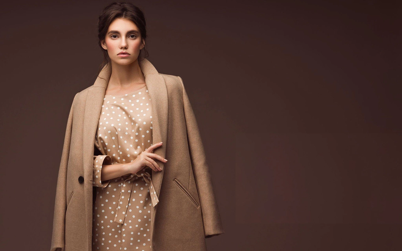 Une femme vêtue d'un manteau beige et d'une robe à pois se dresse sur un fond marron foncé, incarnant la sophistication discrète et l'exclusivité du 'luxe tranquille' s'orienter.