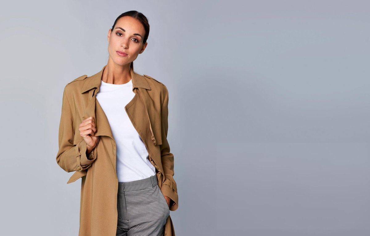 Woman demonstrating practical fashion tips with layered capsule wardrobe outfit featuring camel trench coat over white t-shirt and patterned skirt showing versatile daily styling