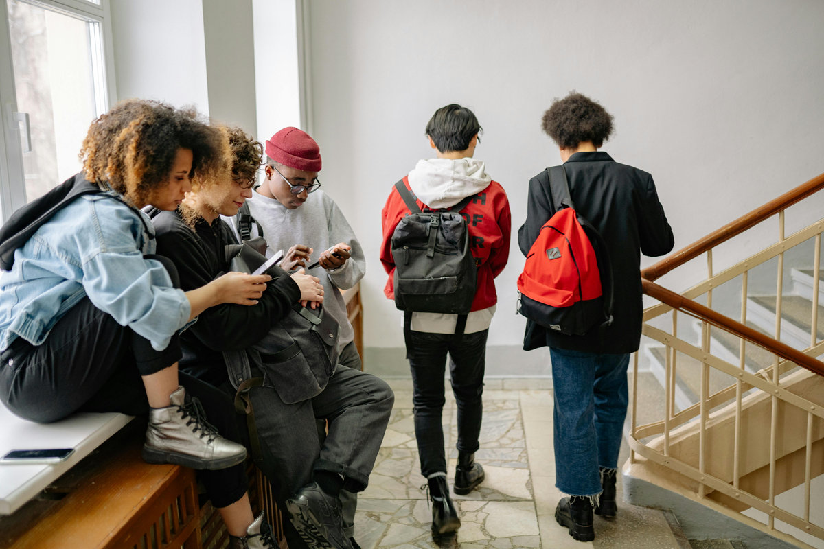 Group of four diverse university students on campus staircase displaying varied fashion styles from casual streetwear with hoodies and denim to semi-formal blazer, carrying backpacks and interacting socially