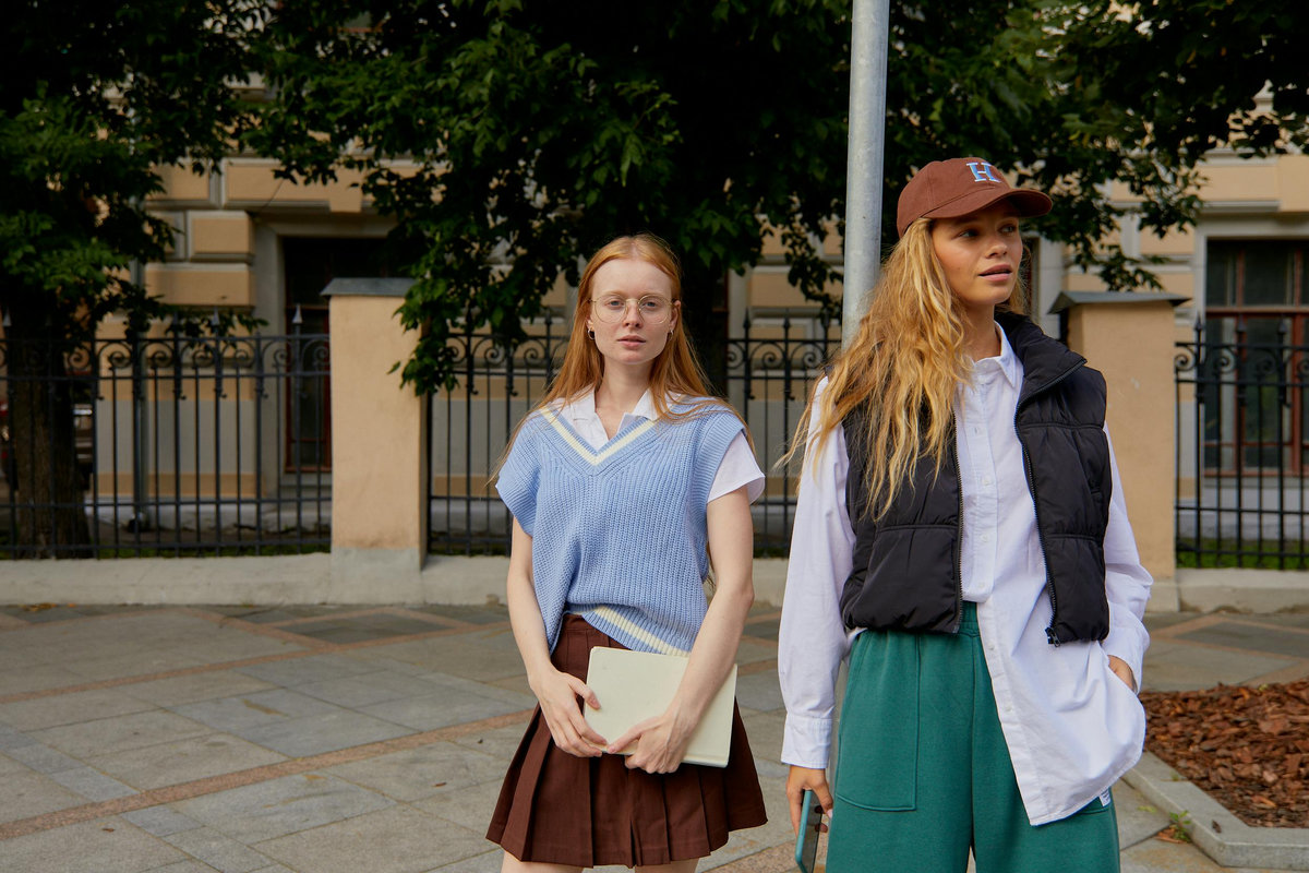 Two female university students outside campus building displaying contrasting fashion styles—preppy academic look with knit vest and pleated skirt versus casual athletic style with puffer vest and baseball cap