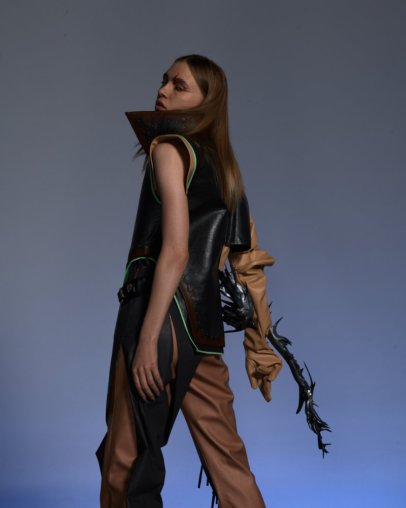 Side profile of model in Xuhui Zhao design featuring black sleeveless top with geometric neckline, tan trousers with black spike embellishments on sleeve