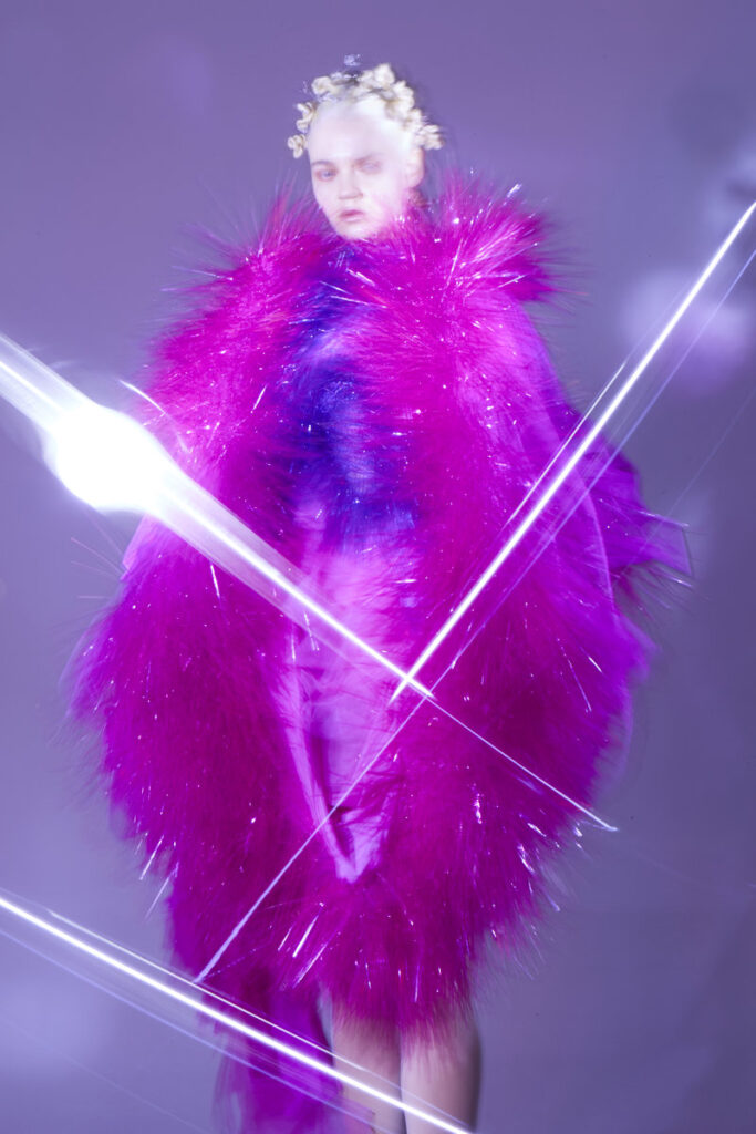 Model enveloped in voluminous magenta feathered coat by Xuhui Zhao with dramatic white light streaks crossing the composition against purple background