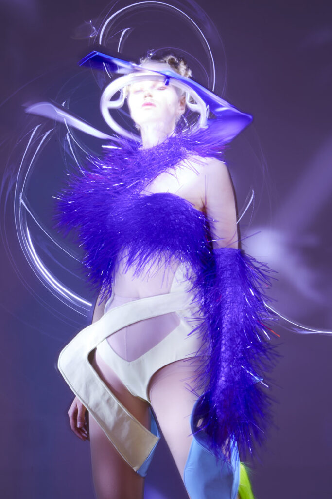 Model in Xuhui Zhao design featuring violet feather embellishments, white sculptural elements, and geometric neon light headpiece with circular light installation