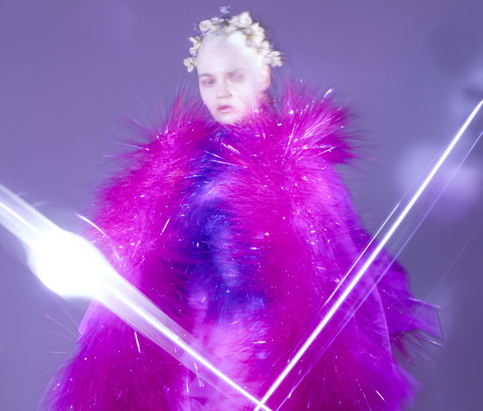 Model enveloped in voluminous magenta feathered coat by Xuhui Zhao with dramatic white light streaks crossing the composition against purple background
