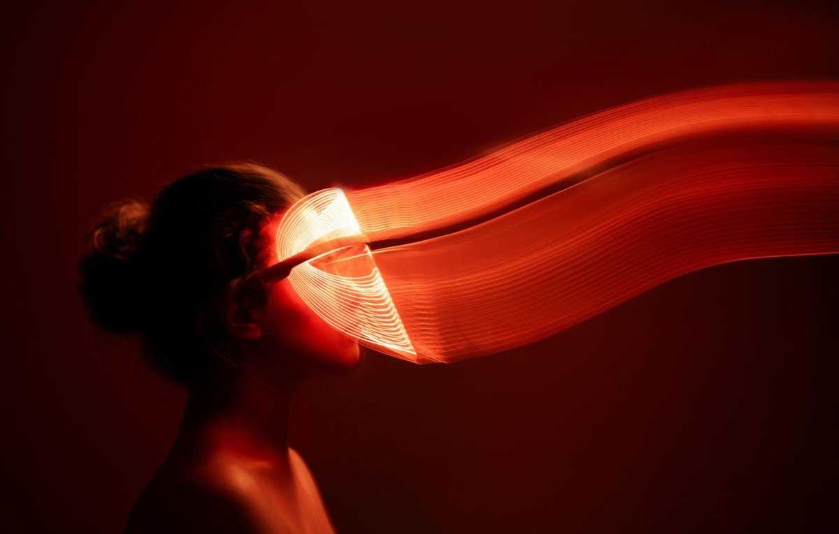 Woman in profile wearing an LED face mask with red light trails against a dark background
