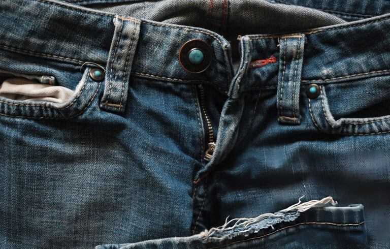 Close-up of worn and fraying denim jeans with turquoise rivets and distressed fabric — The Fashion Globe