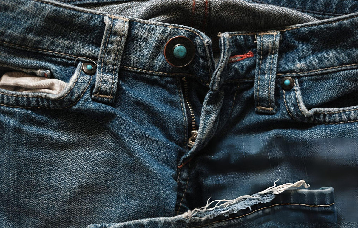 Close-up of worn and fraying denim jeans with turquoise rivets and distressed fabric — The Fashion Globe