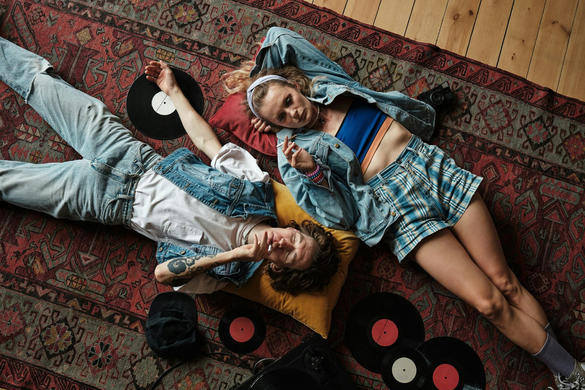 Two people in vintage denim clothing lying on a patterned rug surrounded by vinyl records