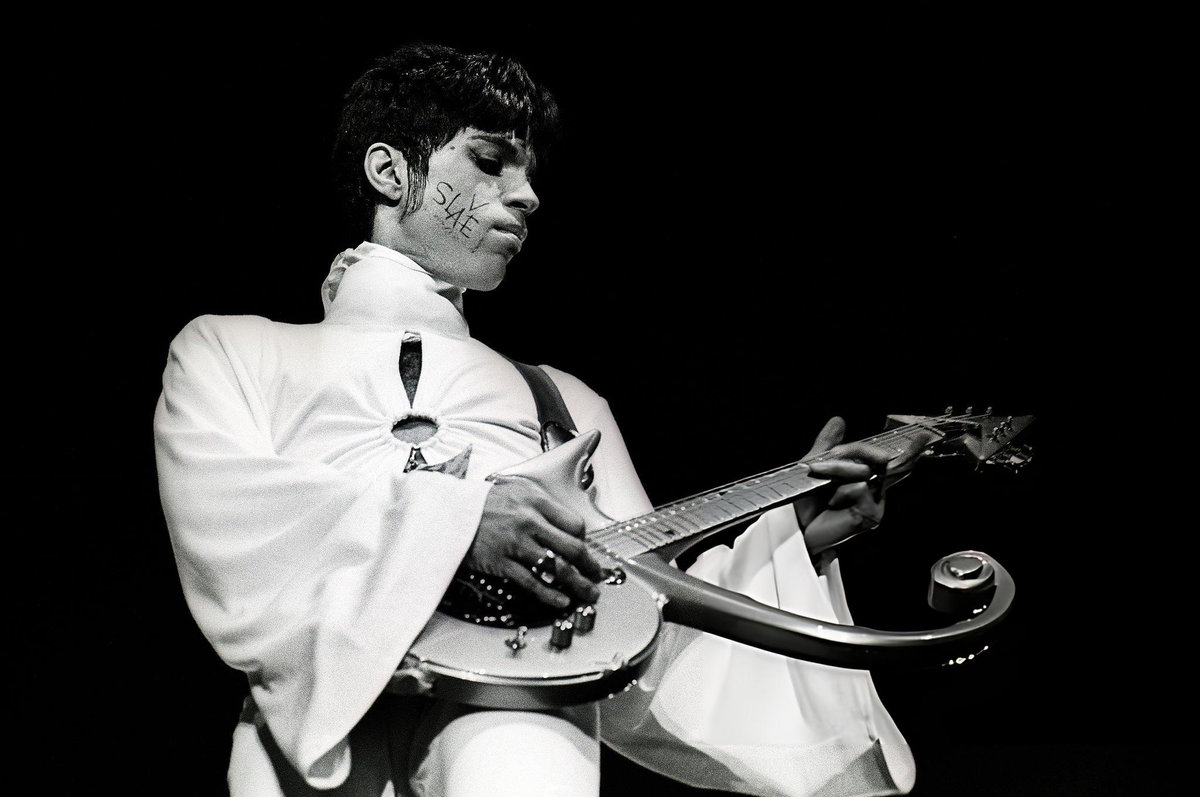 Prince performing on stage in a white suit, the word SLAVE written on his cheek, playing his Love Symbol-shaped guitar at Brabanthallen, Den Bosch, Netherlands, 1995 — The Fashion Globe
