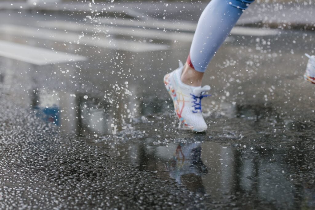 Close-up of runner's feet in sneakers splashing through puddles on a wet street.