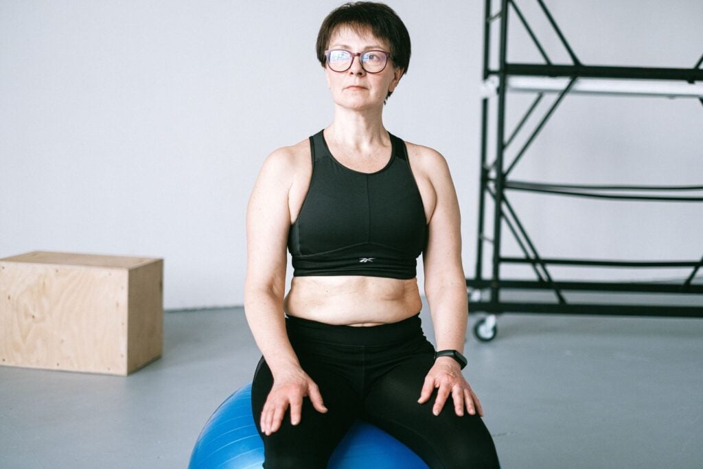 Middle-aged woman with eyeglasses exercising on a blue stability ball indoors.